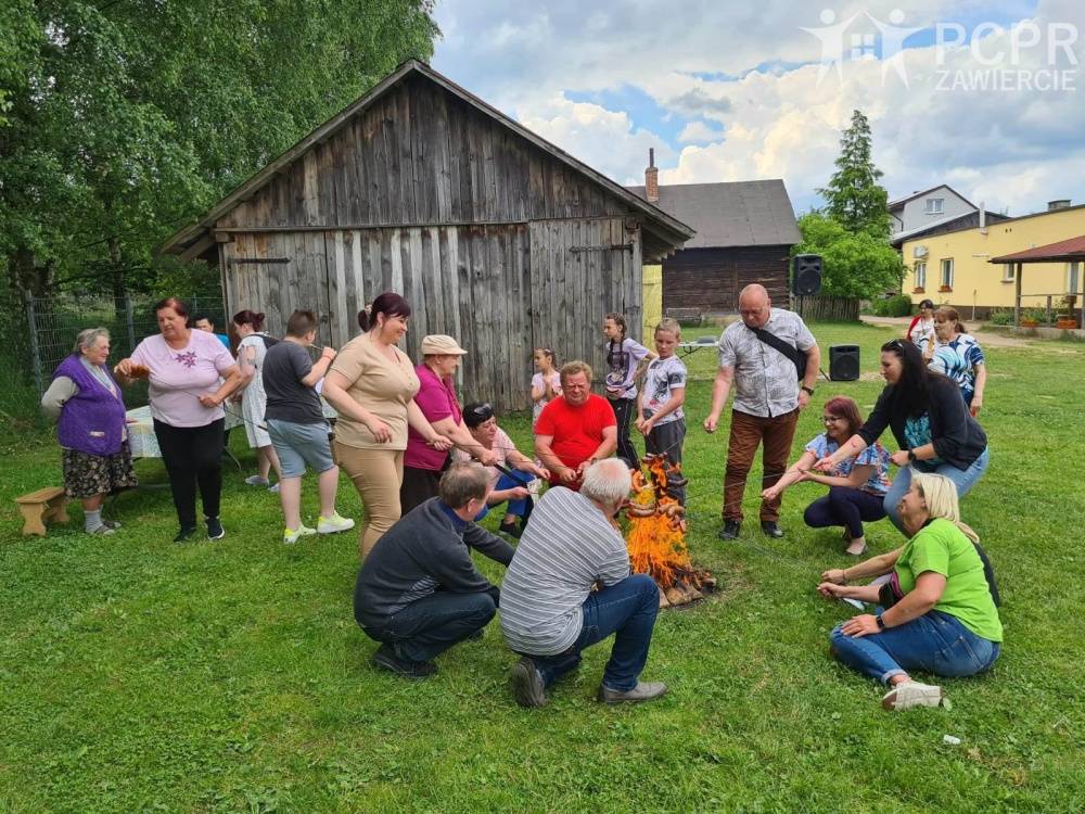 Zdjęcie: Grupa ludzi stoi i siedzi przy ognisku i piecze kiełbaski na patykach. Są zadowoleni.